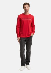 Mann steht mit einer Hand in der Tasche, trägt ein rotes Langarmshirt mit Logo, dunkle Jeans und weiße Sneakers vor einem weißen Hintergrund.