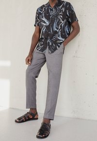 Uomo in pantaloni grigi su misura, camicia a maniche corte con motivo nero e blu, e sandali in pelle nera con punta aperta, in piedi contro un muro bianco.