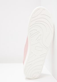 Semelle d'une chaussure rose avec une semelle blanche montrant des motifs de bande de roulement courbés et angulaires, placée sur un fond blanc.