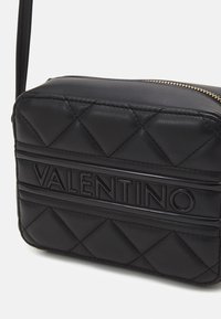 Juodas siuvinėtas odinis perpetėnos krepšys su įspaustu "VALENTINO" logotipu, auksine užtrauktuko detale, stačiakampio formos ir reguliuojama petneša.