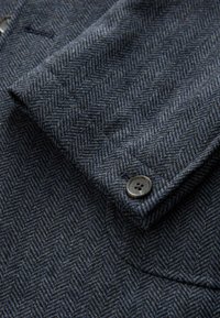 Woll-Herringsbone-Stoff in dunklen Blau- und Grautönen. Verfügt über ein Knopfdetail und eine weiche Textur mit einem gemusterten Design.