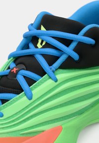 Sapata desportiva com sola verde brilhante, detalhe lateral em verde neon, língua preta e atacadores azul elétrico cruzando na parte superior.
