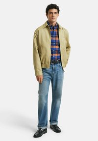 Beige leichte Jacke, kariertes Hemd in Blau und Orange, blaue Jeans, schwarzer Gürtel und schwarze Schuhe. Lässiges Outfit mit verschiedenen Texturen.