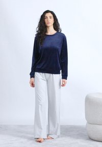 Γυναίκα με μακριά σγουρά μαλλιά που φοράει μπλε ναυτικό μακρυμάνικη μπλούζα και ανοιχτό γκρι παντελόνι με φαρδιά μπατζάκια, όρθια ξυπόλυτη σε χαλιμένη επιφάνεια.