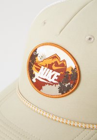 Pălărie bej cu un patch rotund brodat care înfățișează munți, copaci și logo-ul Nike în alb, completată de un accent împletit de culoare portocalie și galbenă.