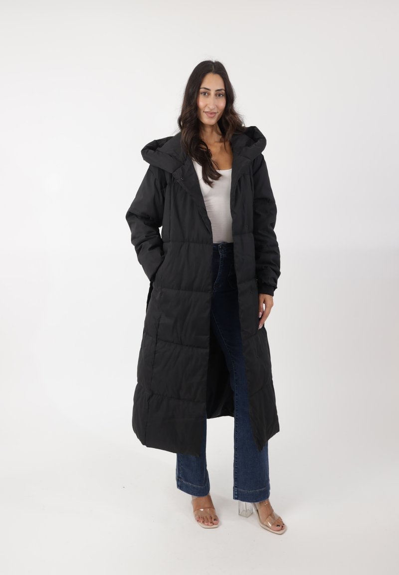 Abrigo acolchado negro con capucha, con un diseño largo y acolchado. Fabricado con un material texturizado. Combinado con vaqueros acampanados y sandalias de tacón transparente.