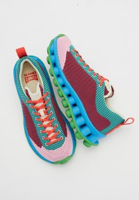 Sapatilha colorida com um cabedal em malha nas cores bordô, teal e rosa, com uma sola espessa e segmentada em azul e verde, e atacadores laranja.