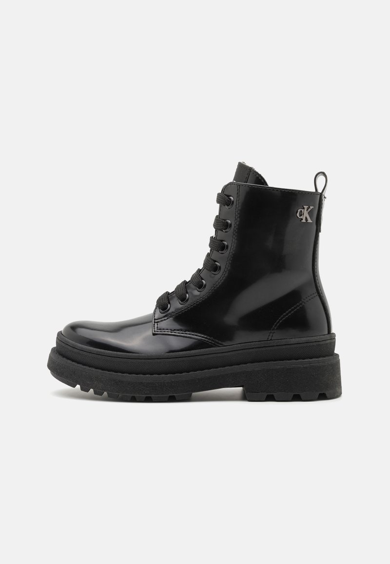 Bottines en cuir verni noir avec une semelle en caoutchouc épaisse, un laçage à l'avant et un accent logo en métal sur le côté. Poids texturé sur la semelle.