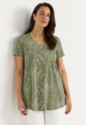 Kobieta z długimi brązowymi włosami, ubrana w zieloną koszulkę z krótkim rękawem i dekoltem w serek z motywem liści oraz białe spodnie, stojąca przy białej ścianie.