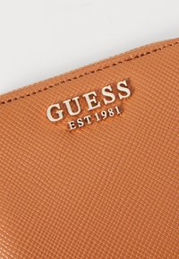 Portefeuille en matériau synthétique texturé, orange, avec motif en relief. Logo argent "GUESS EST 1981" sur le devant. Détail de fermeture éclair sur le bord.