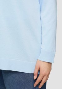 Jasnoniebieski sweter dziany z ściągaczami przy rękawach i u dołu. Zbliżenie na rękaw i dłoń na tle denimu. Gładka faktura.