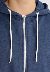 Sudadera con capucha azul marino con cremallera, tela texturizada, cordones blancos y cierre plateado; cuenta con una capucha ajustada y un cuello de canalé.