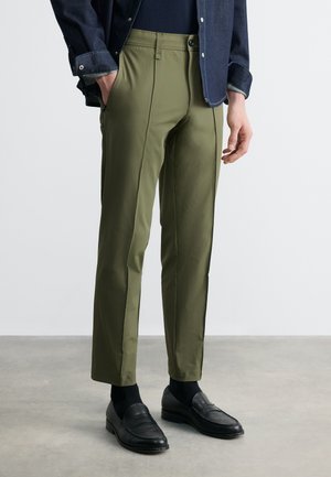 Egy férfi olíva zöld, szabott nadrágot, fekete mokaszint, fekete zoknit és sötét farmerdzsekit visel, szürke padlón áll fehér fal előtt.