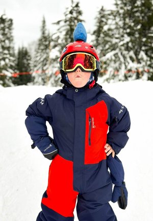Roter und marineblauer Schneeanzug mit Reißverschluss, schwarze Handschuhe und einem roten Helm mit einer blauen Flosse. Große reflektierende Skibrille. Schneebedeckter Hintergrund.