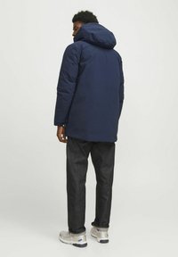 Marinblå puffjacka med huva, med långa ärmar och en avslappnad passform, ihop med mörka jeans och silverfärgade träningsskor.