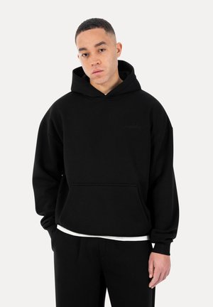 Junger Mann, der einen schwarzen, übergroßen Hoodie und schwarze Hose trägt, vor einem schlichten hellen Hintergrund steht und die Hände in den Taschen hat.