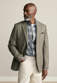 Blazer din tweed în nuanţe de maro, cu model texturat, purtat peste o cămașă albastră în carouri. Pantaloni crem completează ținuta.