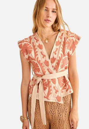 Mujer con top beige cruzado con bordado floral en tono óxido y cinturón a juego, combinado con pantalones marrones de crochet y collar con colgante dorado.
