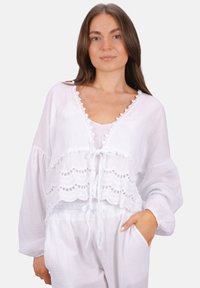 Weißes, kurzes Blusenshirt mit gewelltem Saum und Bindeband, ausgestattet mit langen Ärmeln. Der Stoff ist leicht und strukturiert, kombiniert mit weißen Hosen.