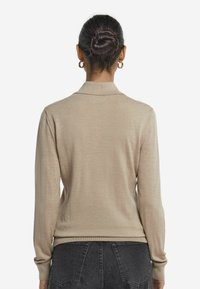 Beige tröja med krage, långa ärmar och ribbade muddar. Mjuk textur, figurnära design, bärs med mörka jeans. Sett bakifrån.