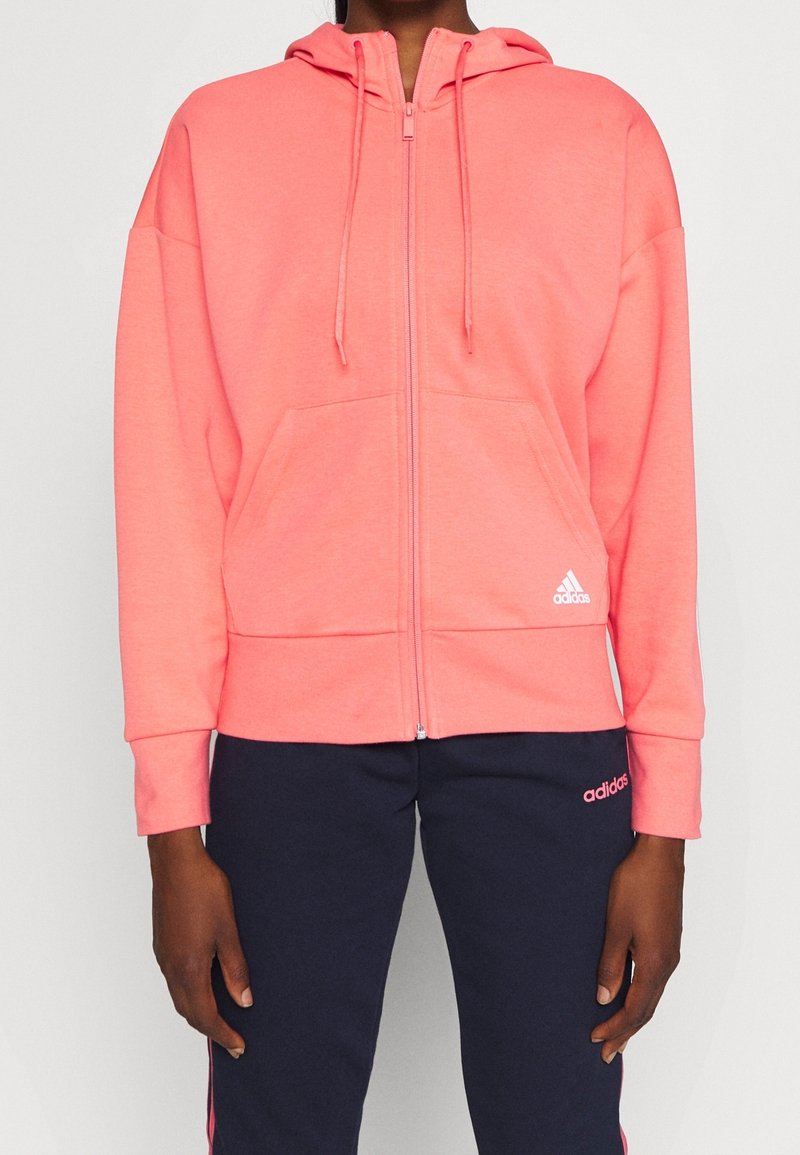Personne portant un sweat à capuche zippé corail rose avec cordons et un pantalon bleu marine, tous deux avec de petits logos Adidas blancs et roses.