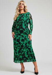 Maximekko vihreänä, jossa on musta kukkakuosi, pitkät läpikuultavat hihat, pyöreä pääntie ja istuva vyötärö. Yhdistetty mustiin korkokenkiin.