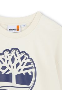 T-shirt color crema con colletto a coste, caratterizzato da un grande logo di un albero blu navy al centro, realizzato in morbido materiale di cotone.