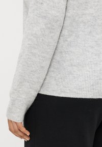 Suéter de malha cinza claro com textura canelada, apresentando mangas longas e um ajuste ligeiramente solto, combinado com calças pretas na cintura.