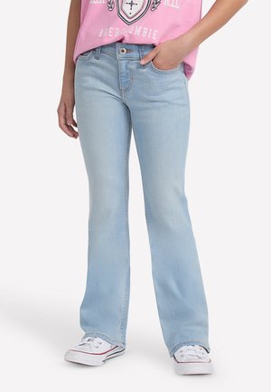 Personne portant un jean bootcut bleu clair, un t-shirt graphique rose rentré, et des baskets blanches avec bordure rouge, une main dans la poche.