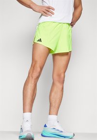 Neon groene sportshorts met zijsplitten, voorzien van het zwarte Adidas-logo. Model draagt witte sneakers met blauwe accenten en witte sokken.