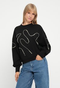 Sweatshirt noir surdimensionné avec des embellissements en perles abstraites. Poignets et ourlet côtelés, avec une coupe ample et décontractée. Assorti à un jean en denim bleu.