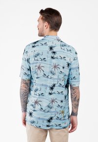 Uomo con tatuaggi su entrambe le braccia che indossa una camicia a maniche corte a stampa tropicale blu chiaro e pantaloni beige, in piedi con la schiena rivolta.