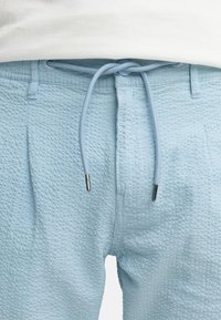 Helleblaue, strukturierte Shorts mit einem Zugband in der Taille, die über metallische Spitzen verfügen. Der Stoff hat ein dezentes, erhabenes Muster für zusätzliche Details.