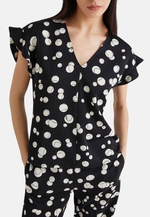 Donna con top a V nero e pantaloni con motivo circolare astratto bianco e maniche corte a sbuffo, mani in tasca.