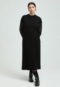 Schwarzes Langarmkleid aus weichem Stoff, mit rundem Ausschnitt, lockerer Passform und kontrastierenden strukturierten Ärmeln. Schwarze Stiefel runden den Look ab.