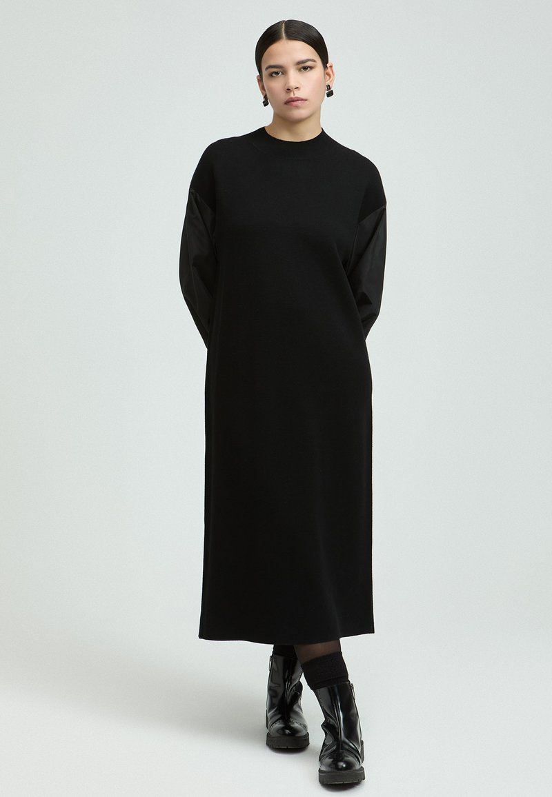 Schwarzes Langarmkleid aus weichem Stoff, mit rundem Ausschnitt, lockerer Passform und kontrastierenden strukturierten Ärmeln. Schwarze Stiefel runden den Look ab.