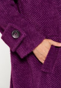 Lila texturerad tygkappa med knäppt manschetter och sidficka. Designen inkluderar en mjuk, vävd yta och skräddarsydda detaljer.