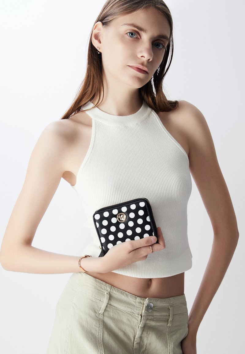 Portafoglio nero a pois con hardware dorato, tenuto da un modello che indossa una canotta bianca a costine, su uno sfondo chiaro.