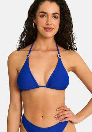Femme aux longs cheveux bouclés portant un haut de bikini halter bleu avec des détails en anneau doré, souriante avec une main sur la hanche.