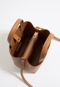 Brązowa skórzana torba-bucket o gładkiej fakturze, ze złotym suwakiem i kieszonką wewnętrzną. Posiada pojedynczy pasek na ramię oraz zaokrąglony górny brzeg.