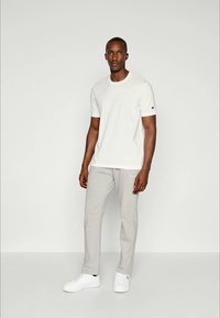 Ein Mann steht in einem weißen Kurzarm-T-Shirt, hellgrauen Jogginghosen und weißen Turnschuhen vor einem schlichten hellen Hintergrund.