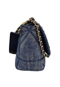 Borsa in denim con design trapuntato, tracolla in catena dorata e un piccolo etichetta attaccata in materiale blu navy. La borsa ha una forma strutturata e arrotondata.