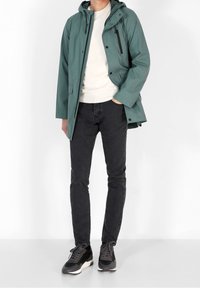 Mężczyzna w turkusowej kurtce przeciwdeszczowej z kapturem, białym swetrze, czarnych dżinsach i czarnych sneakersach stoi na tle białym.