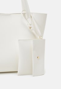 Sac fourre-tout en cuir blanc avec pochette assortie amovible, doté d'un bouton-pression doré et de bretelles d'épaule sur fond blanc.