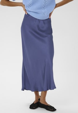 Femme portant un pull en maille bleu et une jupe midi en satin avec des ballerines tissées noires, debout les mains sur les hanches.
