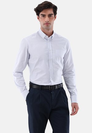 Weiße Hemdbluse mit hellblauen vertikalen Streifen, langen Ärmeln und einer Brusttasche, kombiniert mit navyfarbenen Hosen und einem schwarzen Gürtel.