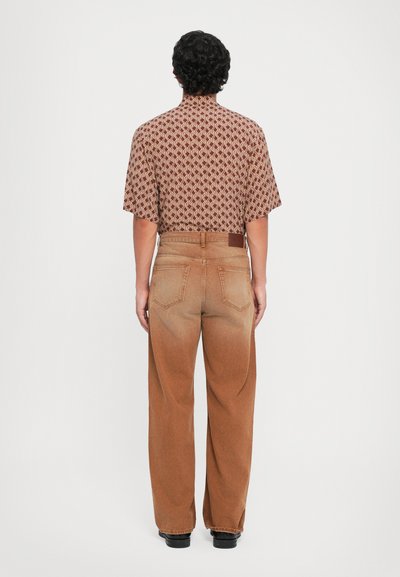 Kortærmet, mønstret skjorte i rød og beige, parret med højtaljede, brede brune denimjeans. Bagudsigt viser lommer og falmede detaljer.