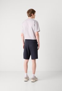 Grå kortärmad skjorta och marinblå shorts. Shortsen har en bakficka; outfiten har en avslappnad passform. Modellen bär ljusa sneakers.