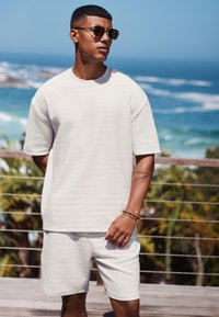 Ljusbeige kortärmad topp och matchande shorts med texturerad design. Accessoarer inkluderar solglasögon och träarmband.