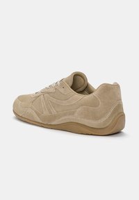 Beige suede sneaker with a textured surface, lace-up design, and curved rubber sole. Features stitching accents and a padded collar.